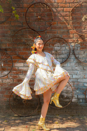 Model in Famameha white printed boho dress with orange accents, flared sleeves, and playful pose against rustic wall, luxury slow fashion from India